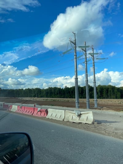 The image shows a row of three high-voltage power transmission towers erected next to a road in Novodrozhzhino, Russia. A blue sky with cumulus clouds forms the backdrop. The towers appear to be newly installed, and power lines are strung between them. Concrete barriers painted red and white line the edge of the road. The road is asphalt, and a forest borders the opposite side of the towers. The view is taken from inside a vehicle, as evidenced by the side-view mirror visible in the lower right corner of the image.