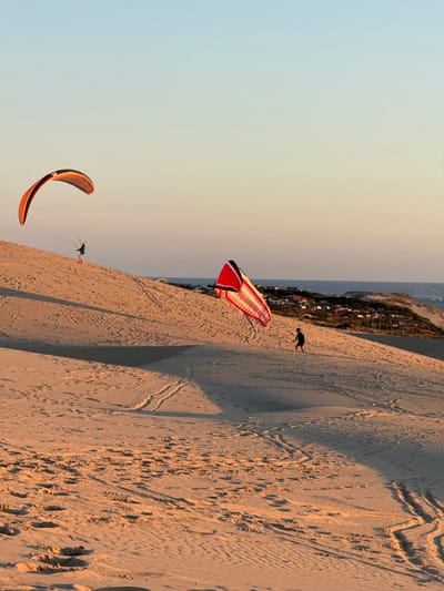 The image shows the Dune of Pilat in La Teste-de-Buch, France, during sunset. Two individuals are visible on the sand dunes, one paragliding and the other walking with a kite. The paraglider, wearing a blue jacket and yellow pants, is suspended in the air with a colorful parachute above them. The walker, dressed in dark clothing, is carrying a large red and white striped kite. The sand dunes are golden in the low sunlight, with footprints and tire tracks visible on the sandy surface. In the background, the Atlantic Ocean stretches to the horizon, with a small cluster of buildings and trees on a coastal ridge. Several other paragliders can be seen in the distance, adding to the sense of activity and recreation in this popular tourist destination.