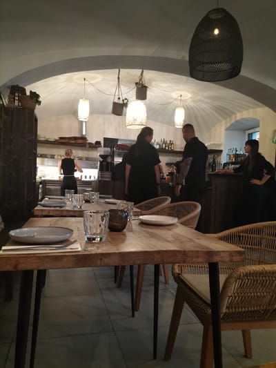 The image depicts the interior of a restaurant, possibly located in Pardubice, Czechia, characterized by its rustic and modern design. The ambiance is warm and inviting, with soft lighting emanating from decorative lamps and accent lighting highlighting the arched ceiling. Several tables are set with plates, cutlery, and glasses, indicating a dining area. A central focus is a wooden table in the foreground, adorned with a light-colored plate, cutlery, and glasses. Two woven chairs with cushioned seats are visible, one on the left and another partially on the right, suggesting a comfortable seating arrangement. In the background, staff members are observed working. A woman with blonde hair is seen behind a counter in what appears to be the kitchen area. Two other individuals, one with tattoos on his arm and another woman, are standing near a bar counter, potentially interacting with customers or preparing drinks. The restaurant's decor includes dark wood elements, shelves stocked with bottles, and potted plants, contributing to a cozy and chic atmosphere.