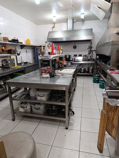 The media file shows a view of a commercial kitchen. The scene is a well-lit, tiled space with stainless steel appliances and work surfaces. Prominently featured is a large stainless steel island table in the foreground, with shelves underneath holding various storage containers and kitchenware. To the left, a stainless steel meat grinder is visible on a shelf, along with stacks of wooden cutting boards and what appear to be plates.
In the background, a commercial-grade ventilation hood is mounted above a cooking area with more stainless steel equipment. Several pots and pans are visible on the shelves and surfaces. Various bottles, likely containing condiments or cleaning supplies, are arranged on a shelf above the island. On the floor, several green cylindrical containers, possibly for waste or ingredients, are placed near the cooking area.
The overall impression is of a functional, working kitchen. The lighting suggests it is daytime or the lights are on. There is no visible activity of people in the image, implying the kitchen is either between uses or captured from a distance. The setting appears to be a professional food service establishment. Visible text includes "GIZARTMALII" and "ARTMALII SUN" on the green containers, and some lettering on the island table and storage bins. The location is identified as Buzovna, Azerbaijan.