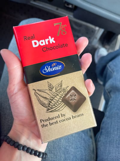 A close-up shows a person's hand holding a rectangular box of "Shoniz" brand dark chocolate. The box has a red, black, and brown color scheme. The red section has "Real Dark Chocolate" and the number "78" printed on it. The black portion contains the brand name, "Shoniz" in a blue oval. The brown section displays a sketch of cocoa beans and a square of chocolate. Text on the brown section states, "Produced by the best cocoa beans." The person is wearing a black beaded bracelet. The background suggests the interior of a vehicle or indoor space, with blue denim clothing and a white sneaker visible. There is no information to suggest the city of Moskovskoe, Russia, as being the location of this image.