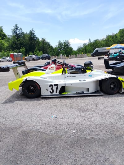 The image shows a race car with the number 37 on its side, parked on an asphalt surface, likely a racetrack. The car is white with yellow accents and has a driver's helmet inside. In the background, there are trees and a cloudy sky. Several other vehicles, including a van and some passenger cars, are visible further back. The scene appears to be outdoors on a sunny day. There is no indication in the image to suggest the location is the city of Rieti, Italy.