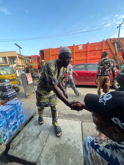 In Akowonjo, Nigeria, a man in a green and blue patterned traditional outfit is shaking hands with another man wearing a black cap and a patterned shirt. The handshake is the central focus, occurring outdoors on a paved surface. Behind them, an orange garbage truck with the letters "W" and "M" visible is parked. A red car is positioned in front of the truck. To the left, a yellow auto-rickshaw and stacks of bottled water are visible, suggesting a market or roadside stall setting. The sky is clear and blue with a few wispy clouds. Other individuals are present in the background, some walking and others seated on a motorcycle. The overall scene depicts a busy, urban environment.