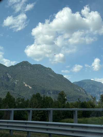The image captures a mountainous landscape under a partly cloudy sky, likely taken from a roadside. In the foreground, a silver metal guardrail with multiple horizontal bars runs across the width of the frame, indicating a road or highway. Beyond the guardrail, a dense line of various green trees forms a buffer before the imposing mountains begin. The mountains are covered in thick vegetation, with some lighter patches of rock visible on their slopes. The sky is a vibrant blue, dotted with fluffy white clouds. A small dark bird is visible in flight high up in the sky. The scene is consistent with the natural beauty of the mountainous regions near Osoppo / Osôf, Italy.