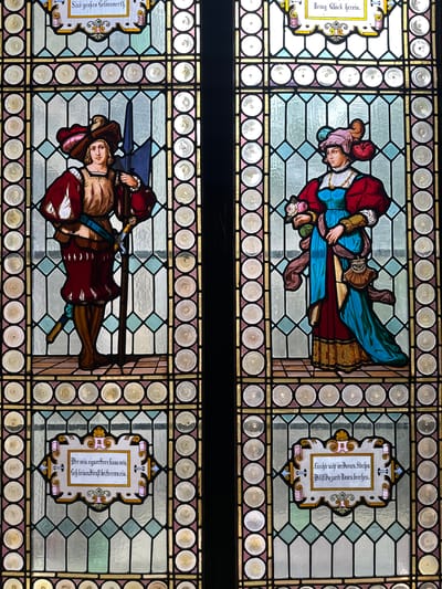 The image shows two stained glass panels, one above the other. The top panel depicts a couple, a man in a red jacket and a woman in a blue dress, embraced. The man is holding flowers. Next to them is a panel with text in an ornate frame. The bottom panel shows a man in armor holding a sword and a banner, with another panel next to him containing text in a similarly styled frame. The stained glass panels are framed by borders of circular glass pieces.