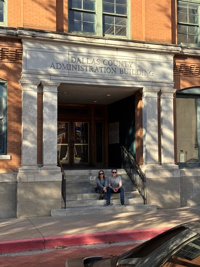 An exterior shot captures the Dallas County Administration Building entrance. The building's façade combines brick and stone architecture. Boldly inscribed above the entrance is "Dallas County Administration Building." A couple, both wearing sunglasses, is seated on the steps leading up to the entrance. A portion of a car is visible on the right side of the image, near the building's edge.