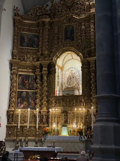 This media file captures the interior of a highly ornate church, specifically focusing on its grand, golden altarpiece, also known as a retablo. The altarpiece dominates the scene with intricate Baroque-style carving, featuring elaborate scrollwork, columns, and decorative elements.
At the center of the altarpiece is a prominent niche housing a statue, likely of a saint or the Virgin Mary, depicted with a radiant sunburst or halo surrounding its head and body. Integrated into the golden structure are several framed paintings, depicting religious scenes with multiple figures in various poses and dark backgrounds.
Below the main altarpiece, an altar is visible, draped in a white cloth. It is adorned with floral arrangements containing orange and white flowers and features a small, illuminated tabernacle or reliquary. Multiple candelabras hold lit white candles, casting a soft glow and contributing to the reverent atmosphere. The candles are placed at different levels on either side of the altar and further up on the altarpiece.
Large, grey stone pillars or columns frame parts of the scene, suggesting the overall architectural style of the church. In the foreground, parts of people's heads and shoulders are visible, indicating the presence of a congregation or visitors attending a service or moment of devotion.
The setting is indoors, with lighting primarily provided by the candles, suggesting either evening or a dimly lit interior. The opulent and detailed craftsmanship of the altarpiece is characteristic of Spanish Baroque art, aligning with the provided location context of San Cristóbal De La Laguna, Spain. There is no discernible text visible in the image.