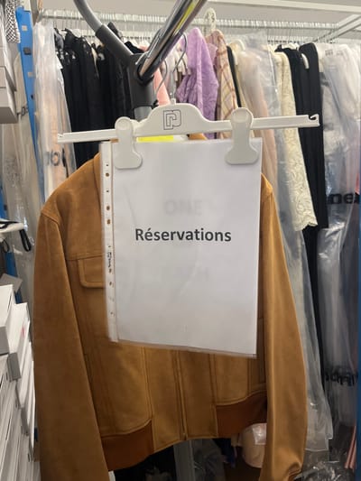 The image, rotated 90 degrees counter-clockwise, depicts a storage or backroom area with numerous items of clothing and organized goods. In the foreground, a prominent brown jacket, likely made of suede or a similar material, is visible. The jacket features ribbed details on what appears to be the cuffs and hem, and has patch pockets on the front.

Attached to the jacket, or hanging in front of it, is a white sheet of paper held within a clear plastic document protector. This plastic sleeve is suspended by a white plastic clip mechanism. The paper clearly displays the word "Réservations" in black, bold, uppercase letters, written vertically from bottom to top (when the image is viewed in its original rotated orientation, it would be horizontally readable). Faintly visible through the paper, possibly on an item behind it or on the jacket itself, are partial letters that seem to spell "ONE WAY M". The plastic sleeve has several punched holes along one side, indicating it's designed for a binder. Small, difficult-to-read text, possibly "Pérez 712", is visible near these holes. A small yellow sticker on the white plastic clip also shows some faint numbers, possibly "2.30".

To the left of the jacket, a silver-colored metal rod or pole is visible, possibly part of a clothing rack, a vacuum cleaner hose, or another piece of equipment.

The background is filled with stacks of various garments. Many clothes are folded and placed on shelves or in bins, some encased in clear plastic bags. A mix of colors is present, including black, cream, and lavender. Further back, more items in clear plastic bags are stacked, with partial text like "per" visible on some of the bags. In the lower right corner of the image, stacks of white boxes are neatly arranged, suggesting organized inventory or packaging. The overall scene implies a systematic storage or preparation area for garments and other goods.