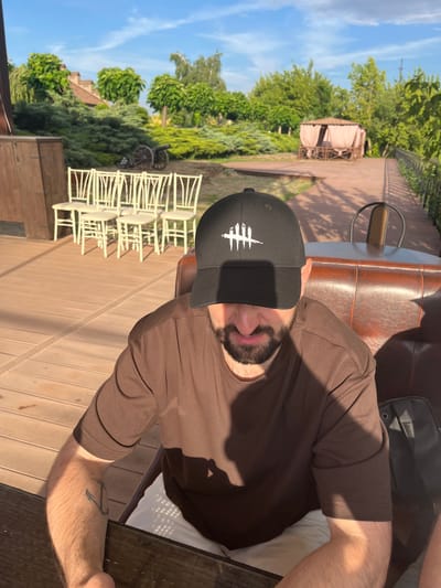 A man wearing a brown t-shirt and a black baseball cap with a white symbol sits on a brown leather couch. He has a beard and is looking at the camera. The scene is outdoors, with a wooden deck in the foreground and lush green trees and a gazebo in the background. Several white chairs are stacked to the left of the frame, and a small cannon is visible behind them. The sun is shining, casting shadows on the deck and the man. The location appears to be a patio or garden area. The overall mood is casual and relaxed. The time of day appears to be late afternoon, given the long shadows. There is no visible text in the image.