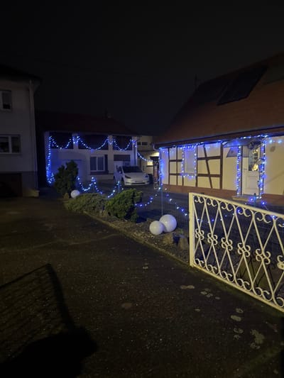 A house is decorated with blue Christmas lights, outlining the roof and windows. Two white spheres sit along the edge of a concrete walkway in front of the house. A white wrought-iron gate is partially visible on the right. A small shrub stands near the spheres. Another house is visible in the background, also adorned with blue lights. The scene is at night, and the sky is dark.