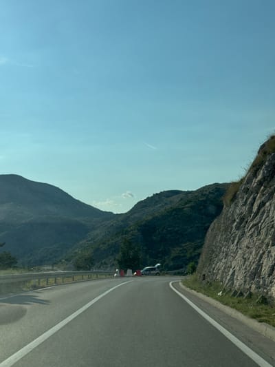 The image depicts a scenic view of a winding road through mountainous terrain, likely in or around Bukovica, Montenegro. The road, paved with asphalt, has a white line marking the center and is bordered by a metal guardrail on the left. On the right, a steep, rocky cliff face rises beside the road, covered in patches of green grass and scrub. In the background, lush green mountains stretch across the horizon, with a clear blue sky above. A few white clouds are visible, and a faint contrail streaks across the sky. Two vehicles are present on the roadside in the distance: a small silver car with its trunk open and a white van. Several orange and white traffic cones are also visible, suggesting roadwork or a temporary closure. The scene is bathed in natural daylight.