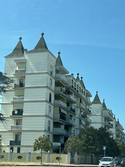 The image captures a row of cream-colored apartment buildings with distinctive turret-like roof structures topped with spheres, set against a clear blue sky. The buildings feature multiple balconies with white railings and striped awnings. In the foreground, a decorative fence with oval cutouts separates the buildings from a grassy area and a street. A street sign indicates "F. Cavallotti," and a traffic sign with a pedestrian symbol is visible. Several trees line the sidewalk. A silver car is parked on the street, and other vehicles are partially visible further down. The scene appears to be a residential street in Montesilvano, Italy.