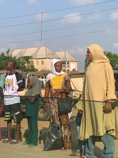 The image depicts an outdoor daytime scene in Abobo, Nigeria, featuring four individuals standing on what appears to be a paved or unpaved street or shoulder.
From left to right:
1. A young male, possibly a teenager, with a shaved head, stands facing left. He wears a white and black raglan t-shirt with graphics, black shorts with white text, and red and black slide sandals.
2. A young female, appearing to be a child, wears a grey hijab covering her head and green long-sleeved top and pants. She stands with her arms crossed, facing left.
3. An adult woman stands centrally, facing slightly right with her gaze directed to the right. She wears a white head covering, a long-sleeved, brightly patterned dress with yellow and brown geometric and swirling designs, and light-colored flip-flops. She carries a black, checkered-patterned handbag on her left arm and a black plastic shopping bag is visible at her feet.
4. Another adult woman stands on the far right, facing left and appearing to be in conversation or looking at the woman next to her. She wears a beige hijab covering her head and a loose-fitting, long beige dress with ruffled cuffs, over blue jeans. She wears light-colored flip-flops.
In the background, behind the individuals, partially visible utility vehicles or trucks are parked. Further back, two residential-style buildings with light-colored, multi-pitched corrugated roofs and light-colored walls are visible under a clear blue sky with scattered clouds. Utility wires traverse the upper portion of the frame, supported by a wooden pole. Some sparse vegetation and ground debris are present on the street level.