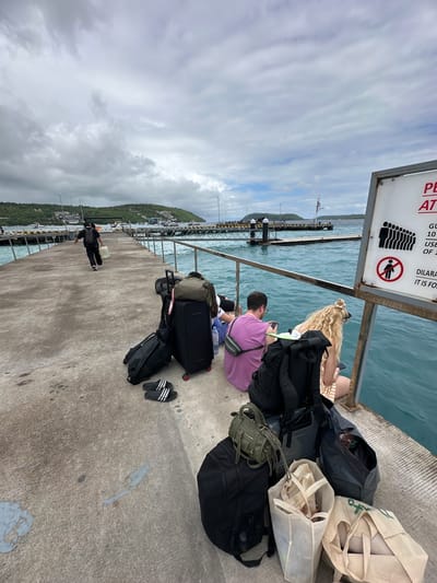 The image shows a group of people waiting on a pier in Kampung Toyapakeh, Indonesia. Several people are sitting near the edge of the pier, by the water. They are surrounded by luggage including backpacks, rolling suitcases, and tote bags. A sign indicating a capacity limit is visible on the left side of the image. A person in black is walking further down the pier. The water and some nearby land are visible in the background under a cloudy sky.