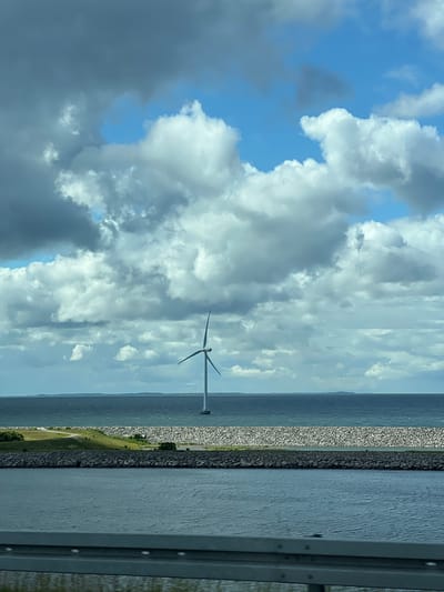 The image captures a view from the Great Belt Bridge in Korsør, Denmark, looking towards the coastline. In the distance, a lone wind turbine stands tall in the calm, blue sea. The foreground shows the water and a section of the bridge's railing. The sky is filled with a mix of dark, dramatic clouds and patches of bright blue. The coastline features a stone embankment, a grassy slope, and a road winding along the shore. The scene is tranquil and showcases a modern wind energy installation against a natural backdrop.