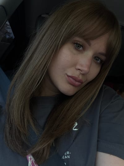 A close-up, slightly angled portrait of a woman with light brown, straight hair and bangs. She has fair skin, visible freckles on her nose, and light-colored eyes. She is wearing a gray t-shirt with a partially visible design on the front. The background is dark, suggesting an indoor setting, potentially a car interior with the edge of a window visible in the lower left. There is no evidence to suggest the location is Shaumyan, Russia. The image focuses on the woman's face and upper torso.