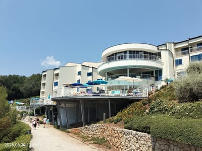 The image shows a white, modern hotel building situated on a hillside overlooking a pathway. The hotel features multiple levels with balconies and large windows, some with blue shutters. A curved section of the hotel faces the viewer, with balconies and white railings. Umbrellas and lounge chairs are visible on the upper decks of the hotel, suggesting a resort or hotel setting. A pathway, made of gravel and lined with low stone walls and vegetation, leads past the hotel. Several people, dressed in casual summer clothing, are walking on the pathway, some carrying bags. The sky is clear and blue, indicating a sunny day. The environment appears to be a coastal area, possibly in Krk, Croatia, given the architecture and vegetation. The foreground features dense green bushes and a stone retaining wall.