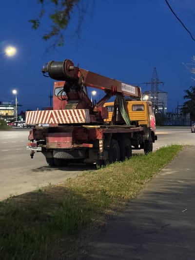 The image shows a yellow crane truck parked on a street in Leninavan, Russia. The crane arm is raised. There are buildings and power lines visible in the background. It appears to be dusk or early evening. Other vehicles are parked further down the street.