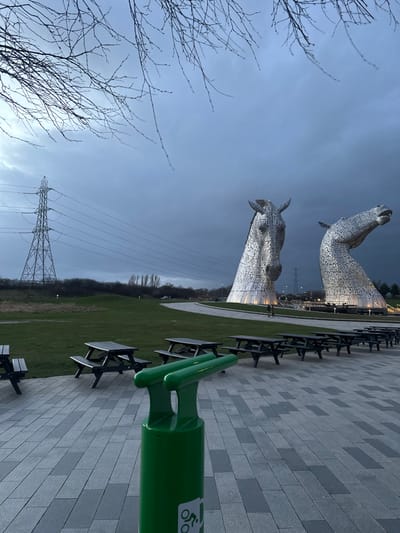 This image depicts The Kelpies, two colossal metallic horse head sculptures, situated in The Helix park in Falkirk, United Kingdom. The sky is dark grey and overcast, with bare tree branches visible in the upper portion of the frame. In the immediate foreground, a bright green cylindrical bicycle repair stand, featuring a white bicycle icon near its base and two upper protrusions, is centrally positioned.
Behind this stand, a series of dark wooden picnic tables with attached benches are arranged on a paved area composed of alternating light and dark grey rectangular slabs. Beyond the paved area, a manicured green grass lawn extends, with a paved pathway curving away to the right. A single, distant human figure, dressed in dark clothing, is walking on this pathway between the picnic tables and the sculptures.
The two Kelpie sculptures are the dominant subjects in the mid-ground, illuminated from their bases. The left sculpture presents a frontal view of a horse's head, while the right sculpture's head is angled upwards. To the far left, a large metal electricity pylon supports multiple power lines that stretch horizontally across the landscape. The distant horizon reveals scattered trees and shrubs. The lighting suggests late afternoon or an overcast day.