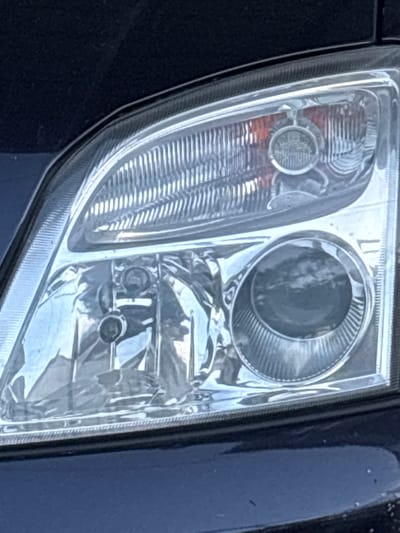 The image shows a close-up view of a car's headlight. The headlight is made of clear plastic with a ribbed interior and has multiple light bulbs visible. A dark blue car body is partially visible around the headlight. There is no indication of the city of Gurkovo, Bulgaria, or any people or actions in the image.