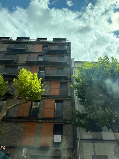 This is a medium shot, eye-level, exterior photograph of a modern brick building with multiple balconies. The building is tall, with several stories visible. The balconies are dark colored with glass railings. Many windows have wooden shutters that are a reddish-brown color and appear to be closed. Two trees with bright green leaves are in the foreground, one on the left and one on the right side of the frame. The sky above is blue with fluffy white clouds. A white towel or sheet is hanging from a balcony on the right side of the building. The bottom of the image shows the facade of the lower part of the building, including what appears to be a sign that reads "ER" in white capital letters on a dark background, and other architectural details. The image appears to have been taken from a street level perspective, possibly from a vehicle, given the slight glare on the left side of the image. The architectural style suggests a contemporary urban setting.