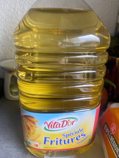 The scene depicts a clear plastic bottle of Vita D'Or "Spéciale Fritures" (Special Frying) cooking oil, holding 2 liters, positioned in an indoor environment in Oberhausbergen, France. The bottle is mostly full of a golden-yellow liquid, featuring horizontal ridges along its transparent body. The product label, primarily light blue with red and green accents, displays "Vita D'Or" in red and green, and "Spéciale Fritures" in blue text. An image of french fries is on the left side of the label, alongside a Nutri-score rating of "C". In the blurred background to the left, a light-colored, possibly white, kitchen scale or similar utensil is partially visible against a textured, light-colored wall. To the right of the bottle, blurred elements include a dark, possibly purple object, a spherical brown object resembling an onion, and the corner of an orange carton labeled "ŒUFS des RÉGIONS" (Eggs from Regions). No people are present, and no actions or interactions are occurring.