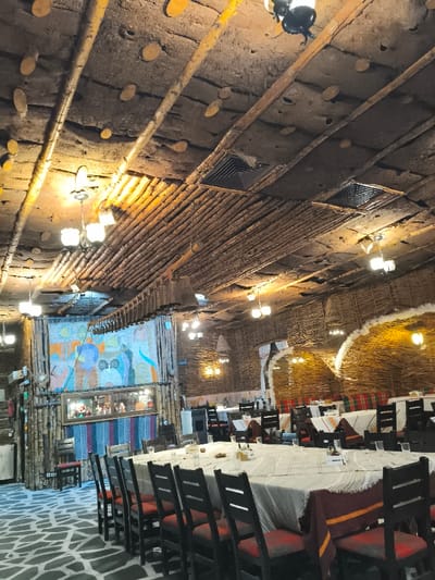 The image depicts the interior of a traditional restaurant or dining hall located in Stoikite, Bulgaria. The perspective is from a slightly elevated position, looking across a large, unoccupied dining area.

In the foreground, a long, rectangular table is prominently set, draped with a white tablecloth topped by a striped fabric runner in red, orange, and brown hues. Dark wooden chairs with red-orange upholstered seats are positioned along both sides of this table. Each place setting includes a clear drinking glass and small condiment dispensers. Similar tables, also set and unoccupied, are visible further into the room.

The ceiling features multiple layers and textures: an upper layer of concrete or plaster embedded with numerous small, round wooden discs, intersected by dark wooden beams. A lower section of the ceiling is constructed from densely packed light-colored wooden branches or logs. Multiple vintage-style pendant lights with frosted glass globes hang from the ceiling, providing warm illumination. Two square, dark ventilation grilles are visible in the ceiling. A long, horizontal decorative element, appearing to be composed of numerous small wooden cylinders, hangs from the ceiling over the main table.

The walls are adorned with various natural materials. The far wall showcases a large, colorful fabric tapestry or mural displaying abstract figures and shapes in tones of blue, orange, green, and white. Below the tapestry, a horizontal mirror reflects indistinct objects. The right-hand walls are predominantly covered in woven branches or wicker, forming large, semi-circular alcoves that are internally lit. Vertical sections of walls are also clad with stacked wooden logs or branches.

The floor exhibits a mosaic-like pattern of irregular grey and white shapes, resembling flagstones. No individuals are present in the scene, and no actions or interactions are depicted.