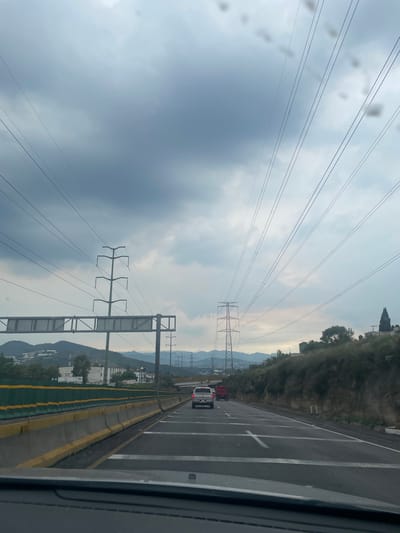 This image captures a highway view in Ciudad López Mateos, Mexico, under an overcast sky. The perspective is from inside a vehicle, looking forward along a multi-lane road. Power lines stretch across the sky, supported by tall, lattice-like towers. On the left side of the highway, a green and yellow barrier separates the lanes from an elevated embankment. Buildings and hills are visible in the distance on the left, while a vegetated hillside with some structures and a tall, dark tree rises on the right. Several vehicles are on the road, including a white pickup truck and a red truck, all moving in the same direction. The road surface is asphalt with white lane markings, including crosswalks at intervals. The overall impression is of a functional, albeit somewhat industrial, urban or suburban transportation corridor.