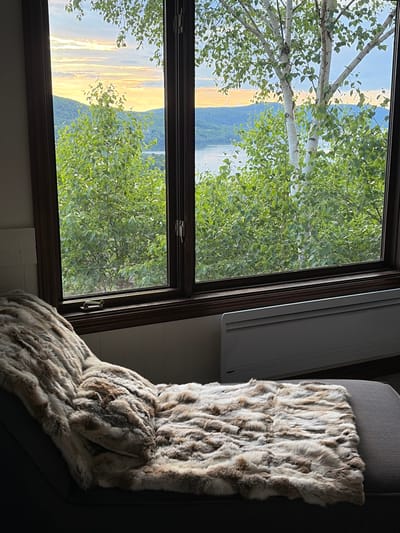 A low-angle, full shot captures an interior view through a large wooden-framed window looking out onto a scenic landscape at dusk. In the foreground, a dark grey chaise lounge is draped with a large, fluffy faux fur blanket featuring a mottled pattern of brown, tan, and cream. The blanket covers most of the lounge, with its texture emphasized by the lighting. Below the window, a long, horizontal, white electric baseboard heater is visible. The window looks out onto a dense forest of green trees, with the trunks of several birch trees prominent, their white bark contrasting with the leaves. Beyond the trees, a wide body of water, likely a lake or river, stretches across the mid-ground, reflecting the soft, warm hues of the setting sun. Distant, rolling green mountains form the background, silhouetted against a sky filled with streaks of orange, yellow, and pale blue clouds. The scene suggests a serene, comfortable setting with a beautiful natural view, possibly in Mont-Tremblant, Quebec, Canada, known for its picturesque mountainous and lakeside environments.
