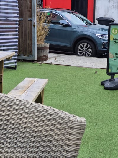 The image depicts an outdoor seating area with artificial turf, located in Carrick-On-Shannon, Ireland. A dark blue SUV is parked on the street bordering the seating area. In the background, a red building and a dark doorway are visible. The foreground shows a wicker chair and a wooden bench. Several small birds are scattered across the artificial turf. A sign with green and white lettering, possibly advertising a garden or shop, is positioned on the right side of the image. A wooden gate and a large potted plant in a barrel are also present. The overall scene suggests a commercial or hospitality outdoor space.