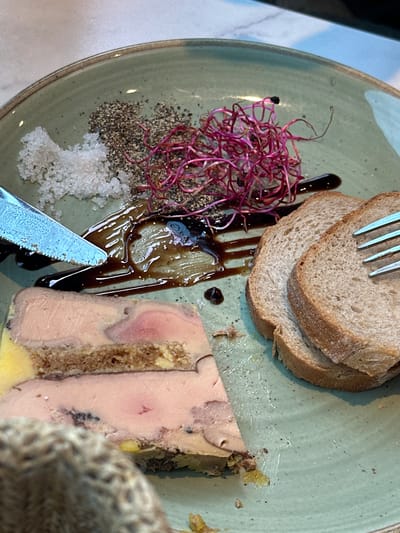 A plate of foie gras is served with toast and garnishes on a light green ceramic plate. The foie gras is a terrine, layered with what appears to be toasted bread or brioche, and is pale pink with a yellow casing. To the left of the foie gras, there is a pile of coarse sea salt and a mound of coarse black pepper. In the center of the plate, a drizzle of dark, glossy sauce, likely balsamic glaze, is spread over a few thin slices of what looks like pickled fruit or vegetable. A bunch of vibrant pink microgreens is scattered over the sauce and pepper. To the right of the plate, three slices of whole wheat toast are stacked. A silver knife with a serrated edge is positioned on the left side of the plate, touching the foie gras and sauce. A silver fork is visible on the right side of the plate, resting on the toast. The setting appears to be an indoor dining environment, possibly a restaurant, with a textured tabletop visible in the background. The scene is illuminated by overhead lighting, creating highlights on the food and plate. This dish is likely from a restaurant in Veules-les-Roses, France, given the context.