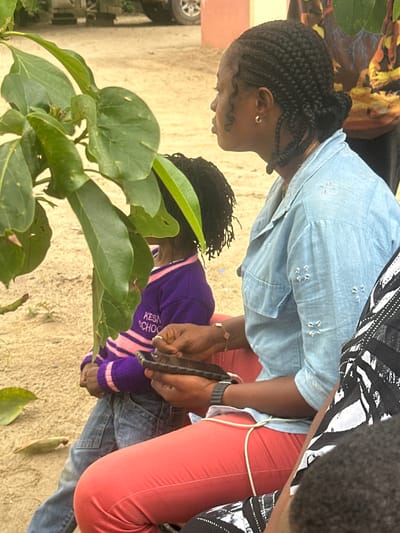 In Ganyingbo, Nigeria, two individuals are seated outdoors on sandy ground near a plant with large green leaves. The person on the right, a woman with dark braided hair, wears a light blue denim shirt and coral-colored pants. She is holding a smartphone in her right hand and appears to be interacting with it. A thin white cord is visible around her waist. To her left, a child with dark hair is visible from the shoulders down. The child is wearing a purple sweater with pink stripes and blue jeans. The child’s hands are not clearly visible. In the background, a portion of a vehicle with a silver wheel is discernible, suggesting an urban or semi-urban setting.