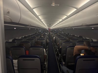 The image depicts the interior of an airplane cabin, viewed from the perspective of a passenger in a rear seat looking forward. The cabin is illuminated by overhead lights, casting a sterile glow on the rows of empty and occupied seats. Passengers are seated on both sides of a central aisle, some looking at personal devices, others resting or conversing. The seats are upholstered in a dark fabric with blue headrests. Luggage compartments are visible overhead. There is no indication of the city of Pitești, Romania, or any other specific location outside of the airplane.