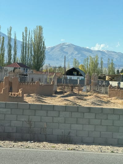 The image shows a view in Toru-Aigyr, Kyrgyzstan. A mountain rises in the background, partially obscured by trees. In the foreground are structures, some in disrepair, including buildings with metal roofs and brick or stone walls. A concrete block wall runs along the bottom right, next to a paved road or pathway. The sky is blue with scattered clouds. The overall setting appears to be a rural or semi-rural area.