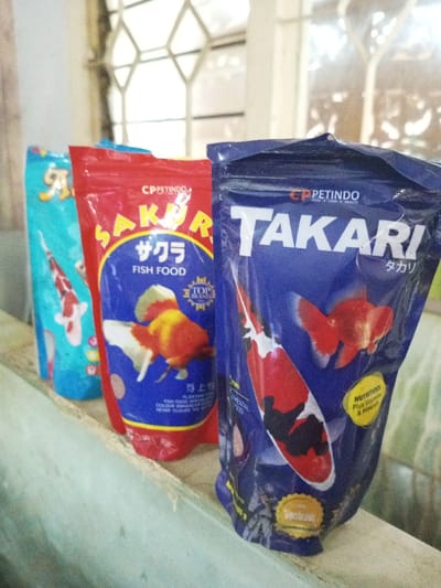 The image shows three bags of fish food lined up on a surface. The bags are labeled "CPPETINDO." The red bag is "SAKURA FISH FOOD" with a picture of a goldfish. The blue bag is "TAKARI" with images of koi fish and goldfish. The cyan bag is mostly out of focus. The fish food packaging has Japanese text, suggesting it's related to koi or ornamental fish. The bags are standing, full, and likely for sale or storage. The brand emphasizes "NUTRITIOUS," "Plus Vitamins & Minerals" and "FOOD CARE & HEALTH."