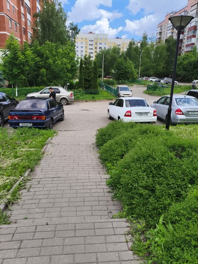 The image depicts a street scene in Malaya Valyaevka, Russia. A gray paved walkway is visible in the foreground, bordered by green grass and dense green bushes. Beyond the walkway, several cars are parked in a lot, including a blue sedan with the license plate T834HK 58, a white sedan with license plate T428TO 58, and a silver sedan with license plate M329EA 5. A man wearing a black t-shirt and a black cap is standing near a silver sedan. In the background, apartment buildings and trees are present. The sky is partly cloudy.