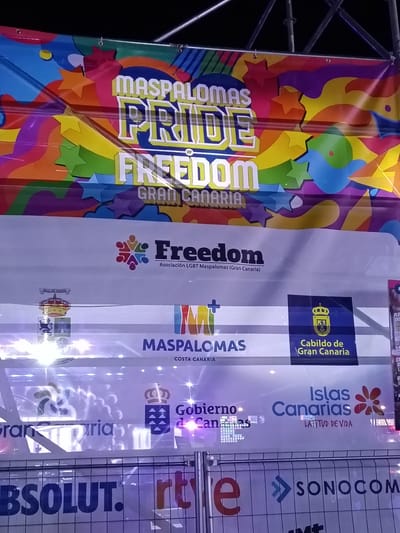 Here is a description of the image:

The image shows a banner for Maspalomas Pride in Gran Canaria, Spain. The banner is colorful and features the words "Maspalomas Pride Freedom Gran Canaria". It also includes logos for various sponsors and organizations, including Freedom Asociación LGBT Maspalomas, Maspalomas Costa Canaria, Cabildo de Gran Canaria, Gobierno de Canarias, and Islas Canarias. Additionally, there is advertising displayed for Absolut vodka, RTVE, and Sonocom. The banner appears to be on a stage or platform. The banner is in the city of Playa Del Inglés, Spain.