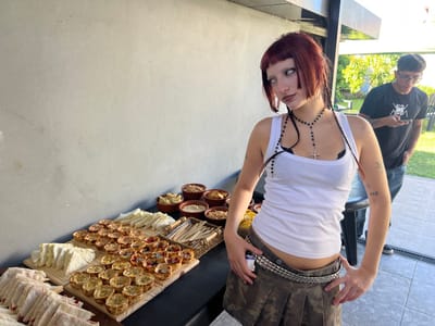 A daytime outdoor scene in Quilmes Oeste, Argentina, depicts two individuals and a food arrangement. In the foreground, a young adult female with dark red hair, styled with blunt bangs and two thin braids, stands slightly angled to the right with her head turned to the left. Her eyes are rolled back, showing white sclera, and she has dark eyeliner and lipstick. She wears a white ribbed tank top, a black bralette, a long dark beaded necklace with a silver cross pendant, a silver-studded belt, and a camouflage-patterned pleated mini-skirt. Tattoos of a small star/asterisk and "222" are visible on her left arm. She maintains a pose with her hands on her hips.
Behind her to the right, partially obscured, a young adult male with short dark hair and glasses stands on a green lawn adjacent to a paved patio. He is observed looking down at a smartphone in his hands. He wears a dark t-shirt with a white graphic design and blue jeans.
A dark-surfaced table positioned in the mid-ground against a light grey wall displays an assortment of food. Items include multiple stacks of triangular sandwiches, some with red filling, arranged on wooden boards. Numerous small savory tarts or quiches are also presented on wooden boards. To the right, several small brown earthenware bowls contain snacks such as chips, nuts, and dips. A woven basket holding wooden utensils is also visible on the table. An architectural beam or overhang is present above the scene.