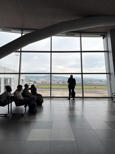 The image depicts the interior of a modern airport terminal waiting area at Matecaña International Airport (Pereira), located in the Area Metropolitana Centro Occidente (AMCO), Colombia. The scene is primarily illuminated by natural light entering through large, floor-to-ceiling windows that offer a panoramic view of an airfield and the distant city of Pereira.
The floor consists of alternating light and dark grey square tiles. Overhead, a slatted ceiling is visible, along with a prominent, curved, light grey structural beam traversing the upper portion of the frame.
On the left side, four individuals are seated in black, metal-framed airport waiting chairs. They are largely silhouetted against the bright exterior. From left to right: an individual with lighter hair is partially visible; a male in a light polo shirt looks downwards, with a dark backpack positioned on the floor in front of him; a male wearing a baseball cap and a dark t-shirt looks towards the right; and another individual, possibly female, with long hair, is seated further back, with a large bag on the floor next to them.
On the right side, a solitary male figure stands facing away from the viewer, looking out the windows. He is dressed in a dark suit jacket and trousers, and a dark, wheeled suitcase rests at his feet to his right.
Through the windows, a section of an airfield runway or taxiway with faint markings is visible in the mid-ground. Beyond the airfield, the urban landscape of Pereira extends across rolling hills, reaching towards a distant mountain range. A white tower structure with antennae is visible through the windows on the far left. The sky is overcast with a consistent grey cloud cover, indicating daytime under cloudy conditions.