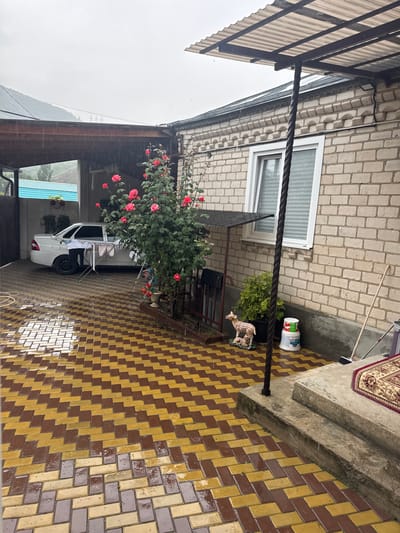 The image depicts an outdoor courtyard in the city of Kamennomostsky, Russia. The ground is paved with alternating yellow and brown bricks, which appear wet, suggesting it is raining. A white sedan is parked under a covered area to the left, with laundry hanging on a drying rack next to it. A dense bush with pink roses is located in the center of the scene. To the right, a white brick building with a window is visible, and a set of concrete steps leads up to an entrance. A decorative statue of a deer with a fawn sits on the patio near a potted plant. A broom and mop are leaning against the wall near the steps. The sky is overcast and gray.