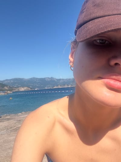 The image captures a partial, close-up perspective of an individual at a coastal location in Kubasi, Montenegro. The subject, positioned on the right side of the frame, wears a brown baseball cap pulled low, obscuring the forehead and casting a significant shadow over the lower face and chest. The visible right eye is dark and directed forward. A golden hoop earring is present on the right ear. The person's right shoulder and upper chest, tanned by sunlight, are visible, with the shadow of the head/cap covering the clavicle area. The background features a clear, bright blue sky above a blue sea. A line of red and white buoys stretches horizontally across the water, marking a boundary. In the distance, dark, rugged mountains form the skyline. On the left side of the frame, a concrete or rocky shoreline abuts the water. Two small figures on an inflatable object are visible in the sea in the mid-distance on the left.