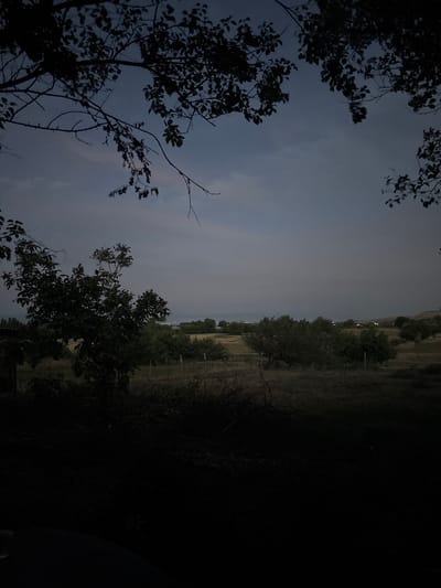 The image depicts a dark, silhouetted landscape with trees in the foreground against a cloudy sky. The scene is likely set in the evening or early morning due to the low light conditions. On the right side of the image, a more detailed view of a rural setting is visible, with rolling hills and scattered vegetation. A fence line can be observed, suggesting a boundary or enclosure within the landscape. In the distance, faint lights can be seen, indicating the presence of human habitation or infrastructure. The overall mood of the image is serene and somewhat mysterious, with the darkness obscuring many details and creating an atmosphere of quietude. The location context provided is Жібек жолы ауылдық округі, Kazakhstan, which is a rural district. This aligns with the visual cues of a natural, somewhat sparsely populated environment. No people or specific objects are clearly discernible due to the darkness, and there is no visible text in the image.