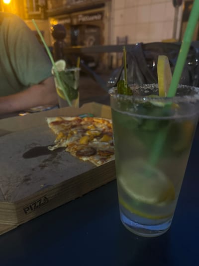 The image depicts a close-up view of a table at night, likely outdoors, with a pizza box and two drinks in the foreground. The pizza box is open, showing a slice of pizza with toppings such as cheese, meat, and possibly chicken or another pale protein. The box has the word "PIZZA" stamped on its side. To the right of the pizza box, there is a clear plastic cup filled with a light-colored beverage, garnished with mint leaves, lemon slices, and a salt-rimmed edge. Two green straws are visible in the drink. The background is blurred but suggests a street scene with illuminated shop fronts, one with signage that appears to read "Patisserie." The overall setting suggests a casual dining or takeaway meal. The specific location in Noyon, France, cannot be determined from the image alone.