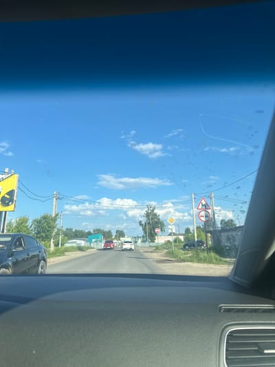 The image shows a view from inside a car, looking out through the windshield at a street scene in Vasilievo, Russia. The sky is blue with scattered white clouds. In the foreground, the dashboard of the car is visible. On the road ahead, there are several cars. A black car is parked on the left side of the road. Traffic signs are visible on the right side of the road, including a warning sign for a winding road and a sign indicating no overtaking. Buildings and trees line the street. A billboard with an image of a black bag and a car part is on the left side of the road. The scene appears to be a sunny day in a semi-urban or suburban area.