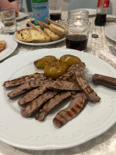 The image shows a close-up of a white plate of sliced grilled steak and small roasted potatoes, served with bread and a glass of red wine on a tablecloth. The steak has visible grill marks and appears to be medium-rare. The potatoes are golden brown and slightly smashed. In the background, a bottle of San Pellegrino water and another glass of red wine are visible, along with a lit candle in a jar. This scene suggests a meal at a restaurant or a dinner setting. Based on the presence of San Pellegrino water and the general ambiance, the location is likely a restaurant in Otranto, Italy, which is known for its culinary scene and scenic coastal views.