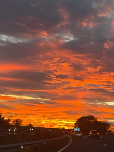 A vibrant sunset dominates the scene with fiery orange and dark gray clouds. The colors transition from intense orange near the horizon to softer hues above. A highway with cars is visible along the right edge. The road curves slightly. Trees line the highway separating it from the sky. The image is taken from a moving vehicle. The composition is dynamic, capturing the fleeting beauty of the sunset and the movement along the road.