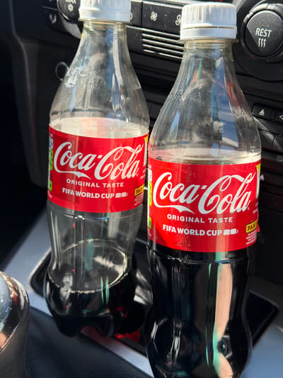 Two bottles of Coca-Cola are visible, one partially empty and the other full, placed in a car's cup holder. The bottle on the left is mostly empty, with a small amount of dark liquid at the bottom. The bottle on the right is full of Coca-Cola. Both bottles have red labels with the Coca-Cola logo and text indicating "Original Taste" and "FIFA World Cup." The surrounding environment suggests the interior of a vehicle, with car controls visible in the background, including a dial labeled "REST" with wavy lines. The image appears to be taken during the daytime, with sunlight illuminating the scene. There are no people visible in the image. The location is Beli Osam, Bulgaria.