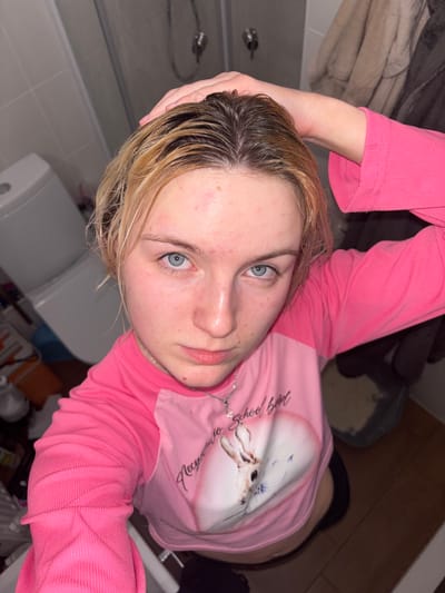 A high-angle, close-up selfie depicts a young person, likely female, with wet hair, dark at the roots and blonde towards the ends. She looks directly into the camera with striking blue eyes and a neutral expression. Her face shows some minor redness or blemishes.
She is wearing a ribbed, long-sleeved pink top. The top features a graphic design on the chest, including a heart shape with an animal (possibly a fawn or deer) inside, and partially visible text that appears to read "My School Beauty." A thin silver chain necklace is visible around her neck. Her left hand is raised, possibly holding or pushing back her wet hair.
The background suggests an indoor setting, likely a bathroom, with light-colored tiled walls and hints of fixtures such as a faucet or shower head on the left and a toilet or sink on the right. The lighting is artificial. The image captures a moment, possibly after washing her hair. The location context provided is Ligolambi, Russia.