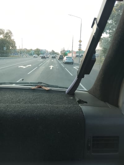 This is a point-of-view shot from inside a vehicle, looking forward through the windshield at a road in Zhlobin, Belarus. The road has multiple lanes and is marked with directional arrows. Several cars are visible on the road, traveling in the same direction. A traffic light is visible in the distance, which appears to be green. On the right side of the road, trees and buildings can be seen. The sky is bright, suggesting it is daytime, possibly late afternoon due to the soft light. The dashboard of the vehicle is visible in the foreground, covered in dark fabric. A decorative element, resembling a bow or ribbon, is placed on the dashboard. The overall scene depicts a normal driving situation on a highway. There is no discernible text visible in the image.