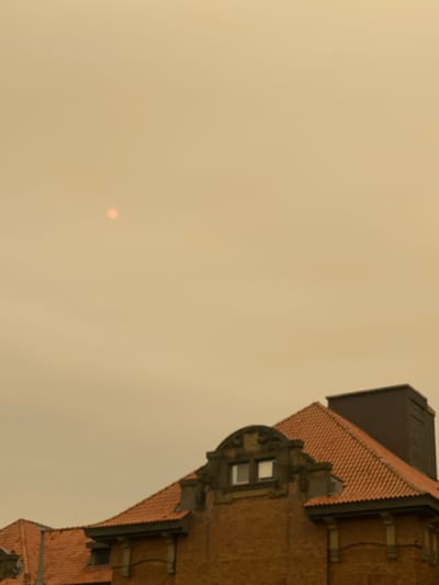 The image shows the upper portion of a brick building with a red tile roof, seen against a hazy, orange sky. A small, reddish orb, likely the sun, is visible in the upper left quadrant of the sky. The hazy conditions suggest the presence of smoke or pollution. The building features architectural details such as decorative stonework above a window and a dark, rectangular structure extending upwards from the roof. The scene appears to be captured from a low angle, looking up towards the building and sky. The specific location in Sainte-Anne-de-Bellevue, Canada cannot be determined from the visual information provided.